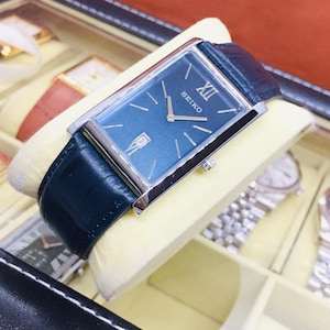 Puede incluir: Un reloj Seiko rectangular con esfera azul oscuro, manecillas plateadas y ventana de fecha. El reloj tiene una caja plateada y una correa de cuero azul oscuro. El reloj se exhibe en una caja de reloj con otros relojes.