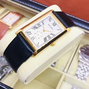 Op de afbeelding: Een goudkleurig rechthoekig Seiko horloge met een witte wijzerplaat en zwarte Romeinse cijfers. Het horloge heeft een zwarte leren band en wordt gepresenteerd op een crèmekleurig kussen. Andere horloges zijn zichtbaar op de achtergrond.