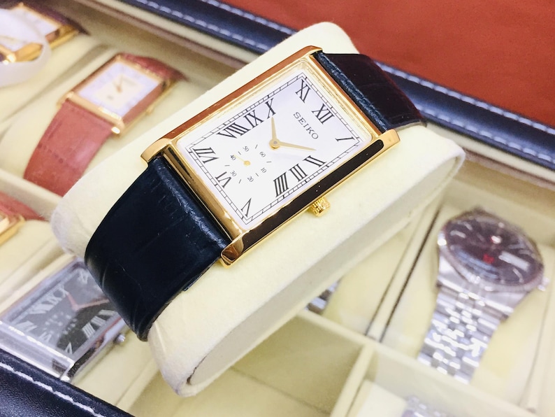 May include: A gold-toned rectangular Seiko watch with a white face and black Roman numerals. The watch has a black leather band and is displayed on a cream-colored cushion. Other watches are visible in the background.