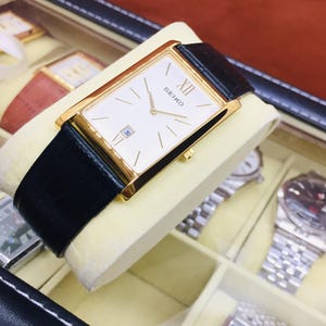 Puede incluir: Un reloj Seiko rectangular dorado con esfera blanca, números romanos y ventanilla de fecha. El reloj tiene una correa de cuero negro y se exhibe en una caja de reloj. La caja del reloj es negra con múltiples compartimentos.