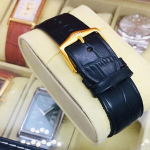 May include: Close-up of a black leather watch strap with a gold buckle. The strap is displayed on a cream-colored cushion inside a watch case. Other watches with various colored straps are visible in the background.