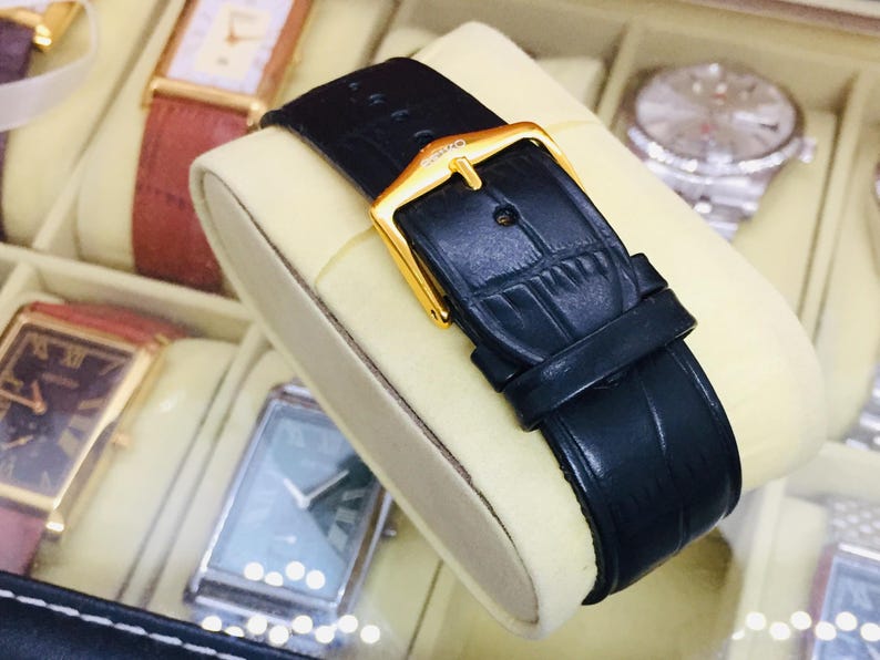 May include: Close-up of a black leather watch strap with a gold buckle. The strap is displayed on a cream-colored cushion inside a watch case. Other watches with various colored straps are visible in the background.