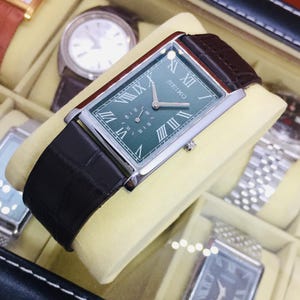 May include: A rectangular Seiko watch with a green face and silver hands, featuring Roman numeral hour markers. The watch has a brown leather strap and a silver case. The watch is displayed in a watch box with other watches.