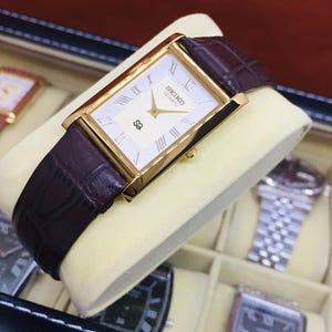 Könnte beinhalten: Eine goldfarbene Seiko Quartz-Uhr mit rechteckigem Zifferblatt und römischen Ziffern. Die Uhr hat ein weißes Zifferblatt und ein dunkelbraunes Lederarmband. Die Uhr wird auf einem cremefarbenen Kissen präsentiert.
