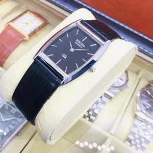 May include: A Seiko Quartz watch with a black rectangular dial and silver case. The watch has a black leather strap and is displayed in a watch case. Other watches with different straps are visible in the background.