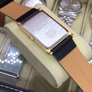 May include: A gold-toned rectangular Seiko watch with a black leather strap. The watch has a silver-colored face and a black leather strap. The back of the watch has the Seiko logo and other text. The watch is displayed in a watch case.