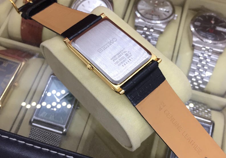 May include: A gold-toned rectangular Seiko watch with a black leather strap. The watch has a silver-colored face and a black leather strap. The back of the watch has the Seiko logo and other text. The watch is displayed in a watch case.
