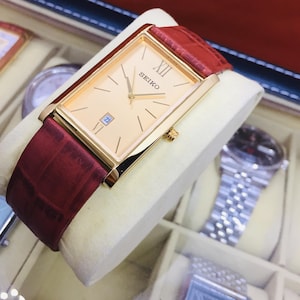 Può includere: Un orologio Seiko dorato con quadrante rettangolare e cinturino in pelle bordeaux. Il quadrante è color champagne chiaro con lancette e indici dorati. La data è visualizzata alle 3. L'orologio è esposto in una scatola.
