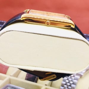 May include: A gold-toned rectangular wristwatch with a black leather band. The watch has a light-colored face and a gold buckle. The watch is displayed on a cream-colored cushion inside a watch case.