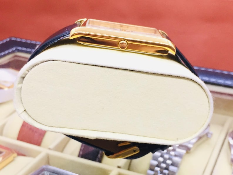 May include: A gold-toned rectangular wristwatch with a black leather band. The watch has a light-colored face and a gold buckle. The watch is displayed on a cream-colored cushion inside a watch case.