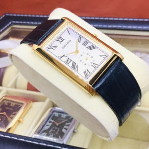 May include: A gold-toned Seiko watch with a rectangular face and black leather band. The watch face features Roman numerals and a small seconds dial. The watch is displayed in a watch box with other watches.