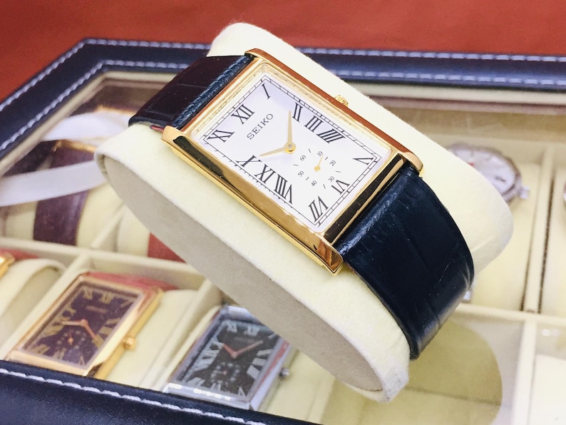 May include: A gold-toned Seiko watch with a rectangular face and black leather band. The watch face features Roman numerals and a small seconds dial. The watch is displayed in a watch box with other watches.