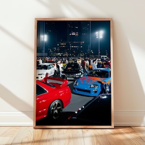 Puede incluir: Una impresión de arte mural vertical enmarcada que representa una reunión nocturna de coches en un aparcamiento. La escena muestra un coche deportivo rojo brillante en primer plano, un coche azul con llamas naranjas y un coche blanco.