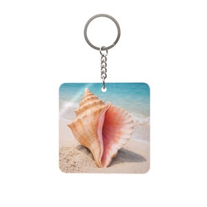 May include: Square keychain featuring a seashell on a sandy beach with a turquoise ocean backdrop. The seashell is a light brown colour with a pink interior. The keychain has a silver-coloured ring and chain.