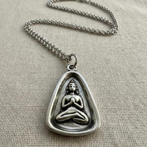 May include: A silver-toned pendant necklace featuring a seated Buddha figure in a triangular frame. The Buddha is depicted in a meditative pose with hands clasped. The necklace has a delicate chain.