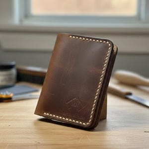 Peut inclure: Un portefeuille en cuir marron avec des coutures crème. Le portefeuille porte un logo et du texte sur le devant. Il est posé verticalement sur une surface en bois. L'arrière-plan comprend des outils et une fenêtre.