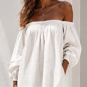 Puede incluir: Un vestido blanco largo hasta el suelo con hombros descubiertos, mangas largas y holgadas con puños fruncidos. La prenda tiene una silueta relajada y fluida con escote fruncido y bolsillos laterales, combinado con un bolso de paja trenzada y sandalias marrones.