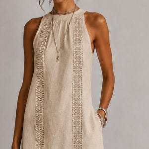 Puede incluir: Vestido largo de lino beige con cuello halter y aberturas laterales. El vestido presenta intrincados paneles bordados en la parte delantera y un bolso tote tejido a juego. La persona lleva sandalias y joyas marrones.