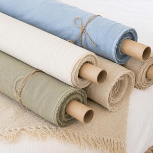 Puede incluir: Cinco rollos de tela en varios colores, incluyendo azul claro, blanco roto, verde salvia y beige natural. Cada rollo est&aacute; envuelto alrededor de un tubo de cart&oacute;n marr&oacute;n y asegurado con cordel fino. La tela tiene una textura tejida visible.