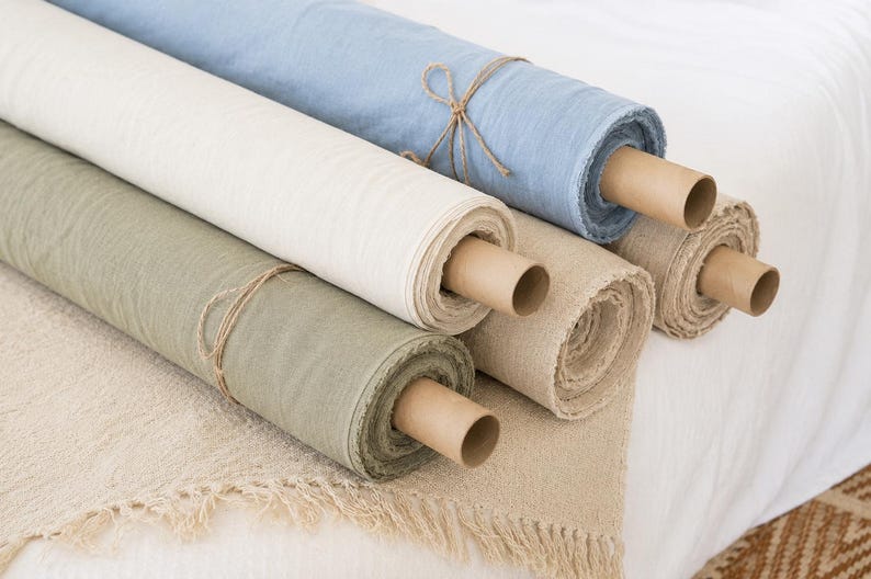 Puede incluir: Cinco rollos de tela en varios colores, incluyendo azul claro, blanco roto, verde salvia y beige natural. Cada rollo est&aacute; envuelto alrededor de un tubo de cart&oacute;n marr&oacute;n y asegurado con cordel fino. La tela tiene una textura tejida visible.