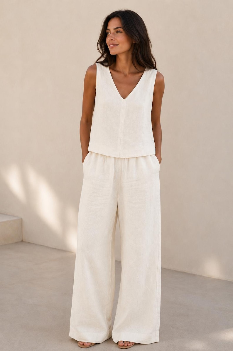 Puede incluir: Un conjunto de dos piezas compuesto por una blusa sin mangas con cuello en V y pantalones anchos a juego en color blanco roto. La blusa presenta un cierre frontal de botones, mientras que los pantalones tienen un corte relajado con cintura el&aacute;stica.