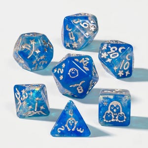 Könnte beinhalten: Set aus sieben blauen und silbernen polyedrischen Würfeln. Jeder Würfel hat silberne Zahlen und Symbole. Die Würfel haben ein durchscheinendes, marmoriertes Aussehen mit Glitzer. Ein Würfel hat ein Pinguin-Design.