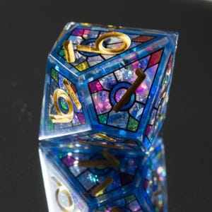 May include: A white ceramic mug with a black handle and the text "But First, Coffee" in a bold, black font. The mug is set against a blurred background, suggesting a cozy setting.