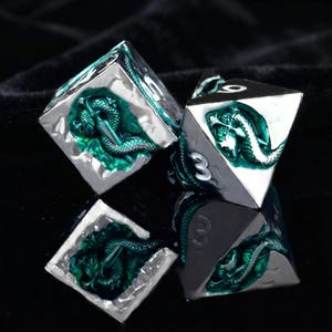 May include: Four polyhedral dice with a silver finish and teal green dragon or serpent design. The dice have white numbering and are displayed on a dark surface. The dice are for tabletop games.