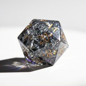 May include: A clear, twenty-sided die with a cracked, iridescent interior. Gold-colored numbers are visible on each face. The die casts a rainbow shadow on a white surface, suitable for tabletop games.