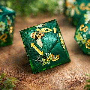 Peut inclure: Un ensemble de dés verts à plusieurs faces avec des chiffres dorés et des motifs d'abeilles et de fleurs. Les dés sont posés sur une surface en bois, avec d'autres dés visibles en arrière-plan. Un objet unique et décoratif.