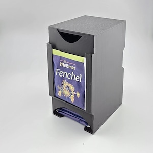 Könnte beinhalten: Ein schwarzer Teebeutelspender mit vertikaler, rechteckiger Struktur und einem offenen Frontschlitz. Ein einzelner Meßmer Fenchel Teebeutel befindet sich in der vorderen Halterung und zeigt eine dunkelblaue Verpackung mit weißem Text und einer gelben Fenchel-Abbildung.