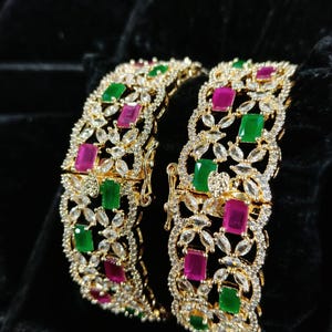 Puede incluir: Dos brazaletes dorados con un diseño floral. Cada brazalete presenta piedras preciosas rectangulares verdes y magenta alternadas, rodeadas de acentos claros y brillantes. Los brazaletes están sobre un fondo negro.