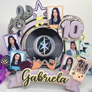 May include: A multi-layered cake topper featuring a central black vinyl record, a large number 10, and the name Gabriela in cursive script. The design includes several small rectangular photo frames, a microphone, a rose, and star shapes.