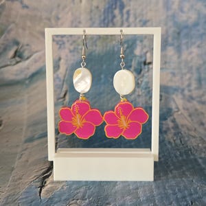 Może przedstawiać: Kolczyki wiszące z wisiorkami w kształcie jasnoróżowych kwiatów hibiskusa z żółtymi środkami i białymi, owalnymi akcentami. Kolczyki są prezentowane na białym stojaku na niebiesko-białym tle.