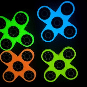 May include: Four glowing fidget spinners in neon green, blue, orange, and lime green against a black background. Each spinner has a central bearing and five arms, creating an X shape. The colors are vibrant and eye-catching.