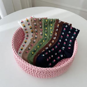 Può includere: Un cestino rosa intrecciato contiene sei paia di calzini fantasia. I calzini sono in vari colori, tra cui bianco, beige, verde, marrone e blu navy, ognuno decorato con piccoli disegni floreali. Il cestino è su una superficie bianca.