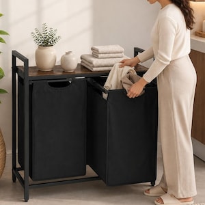 May include: A double laundry sorter featuring a black metal frame and two removable black fabric bags. The unit includes a dark brown slatted wooden top shelf, which holds a small potted green plant, a white ceramic vase, and folded towels.