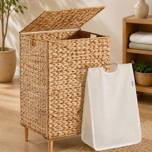 May include: A rectangular woven seagrass laundry hamper with a hinged lid and four light-colored wooden legs. A removable white fabric liner with a metal frame handle is positioned in front of the hamper, showing a clean, neutral aesthetic.