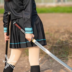 Pode incluir: Uma katana com cabo preto e lâmina prateada. A espada é segurada em frente a uma pessoa vestindo um uniforme preto com detalhes em verde-azulado. A imagem inclui o texto "Katana Forge Legacy".