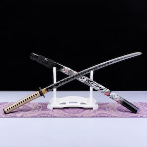 Pode incluir: Uma katana com cabo bege e lâmina preta com padrão em ziguezague, sobre um suporte branco. A bainha apresenta um design gráfico detalhado em preto e branco com detalhes em vermelho, retratando uma criatura e uma lua crescente.
