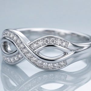 May include: A silver-colored ring with an infinity symbol design. The ring is encrusted with small, sparkling gemstones. The band has a polished finish, and the ring is displayed on a reflective surface.
