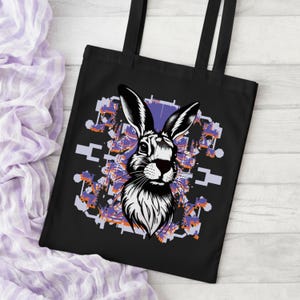 Peut inclure: Sac fourre-tout noir avec une illustration en noir et blanc d'un lapin. Le design comprend un fond géométrique avec des accents violets, orange et blancs. Le sac a des poignées noires et est fait d'un matériau résistant.