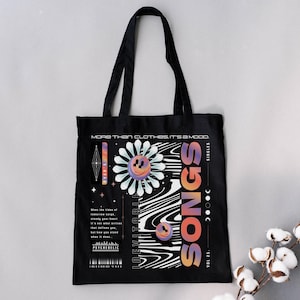 Peut inclure: Sac fourre-tout noir avec un motif graphique représentant une marguerite avec un smiley, le mot "SONGS" en dégradé orange et le texte "MORE THAN CLOTHES, IT'S A MOOD." Le sac a des anses noires et un design minimaliste.