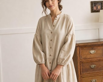 May include: A beige button-down dress with a mandarin collar, long sleeves featuring gathered cuffs, and a loose, flowing skirt. The dress is made of a textured fabric and is paired with a woven straw tote bag held in front of the garment.