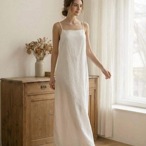May include: A floor-length, sleeveless white dress with a square neckline and thin spaghetti straps. The garment features a straight, relaxed silhouette that falls to the ankles, crafted from a solid, light-colored fabric with a subtle texture.