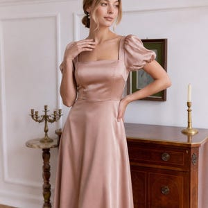 May include: A dusty rose satin midi dress featuring a square neckline, short puff sleeves, and a fitted bodice that transitions into a flowing A-line skirt. The fabric has a smooth, reflective finish, and the garment is constructed with clean seams.