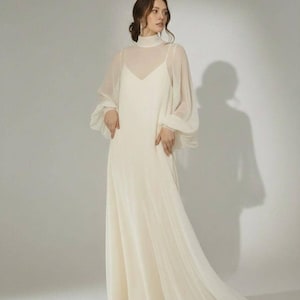 May include: A floor-length cream-colored dress featuring a sleeveless slip-style bodice layered under a sheer high-neck overlay. The overlay includes long, voluminous bishop sleeves with fitted cuffs, creating a soft, flowing silhouette.