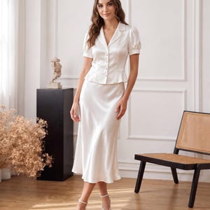 May include: A two-piece set in cream satin consisting of a short-sleeved button-down top with a notched collar and a peplum waist, paired with a matching midi-length skirt that features a smooth, flowing silhouette and a simple hemline.