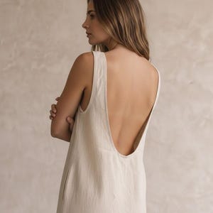 May include: A sleeveless, off-white dress featuring a deep, U-shaped scoop back neckline and wide shoulder straps. The garment is constructed from a textured, light-coloured fabric and falls in a straight, relaxed silhouette down the body.