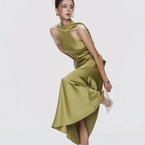 May include: A sleeveless olive green satin midi dress featuring a high neckline with a draped collar detail, a fitted bodice, and a flared skirt that falls below the knees. The garment is paired with white pointed-toe high-heeled pumps and gold jewelry.